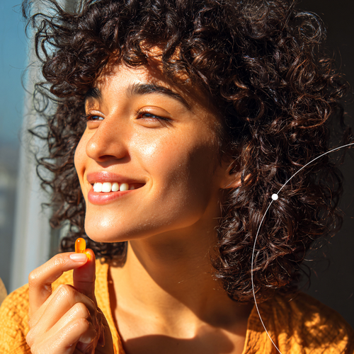 Woman with curly hair smiling outdoors, holding about to take a skincare supplement 